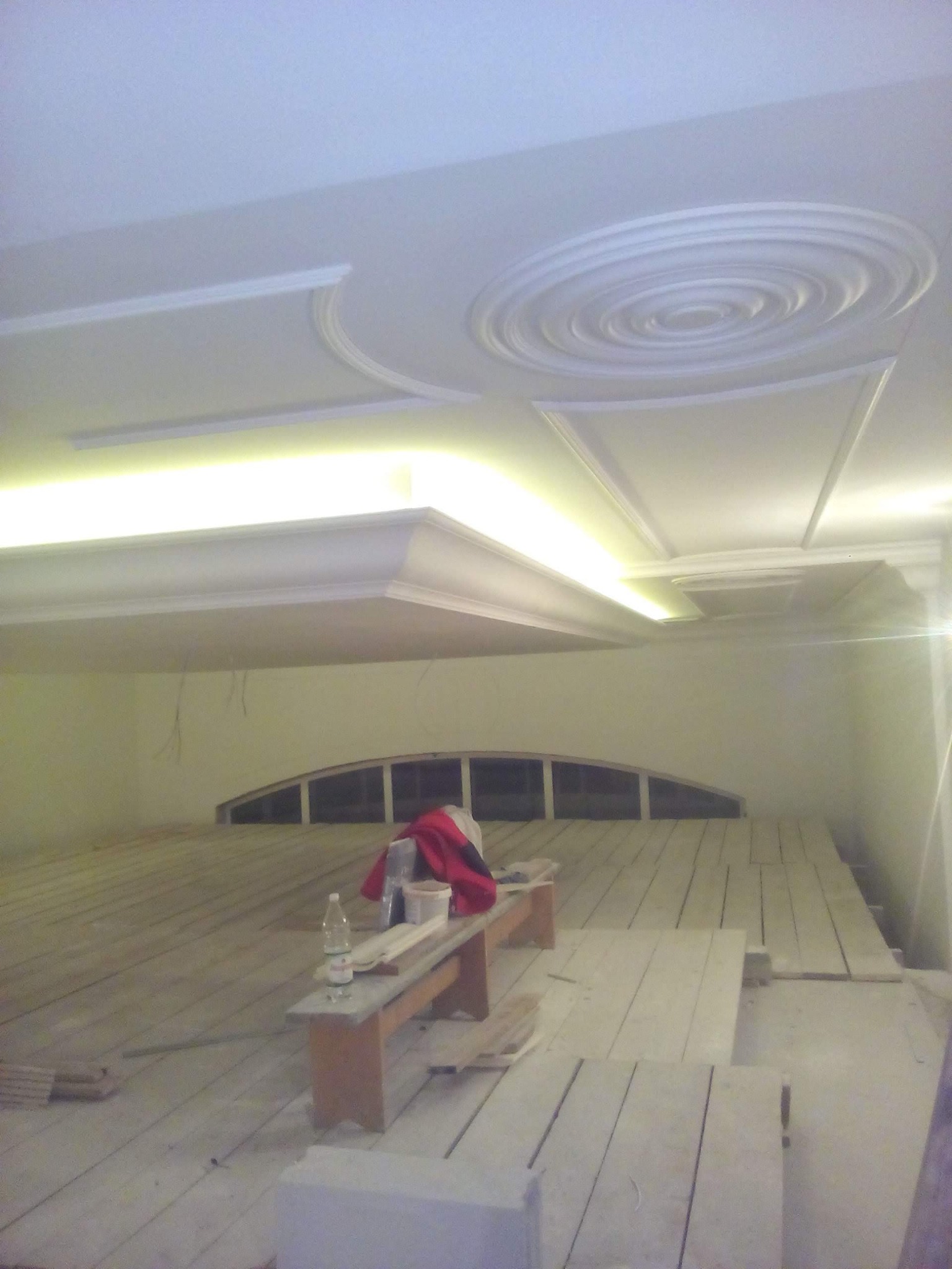 Wnętrze w trakcie prac wykończeniowych: sufit z dekoracyjnymi elementami gipsowymi i podświetleniem LED, częściowo ułożona podłoga z desek, widoczne materiały budowlane i narzędzia.