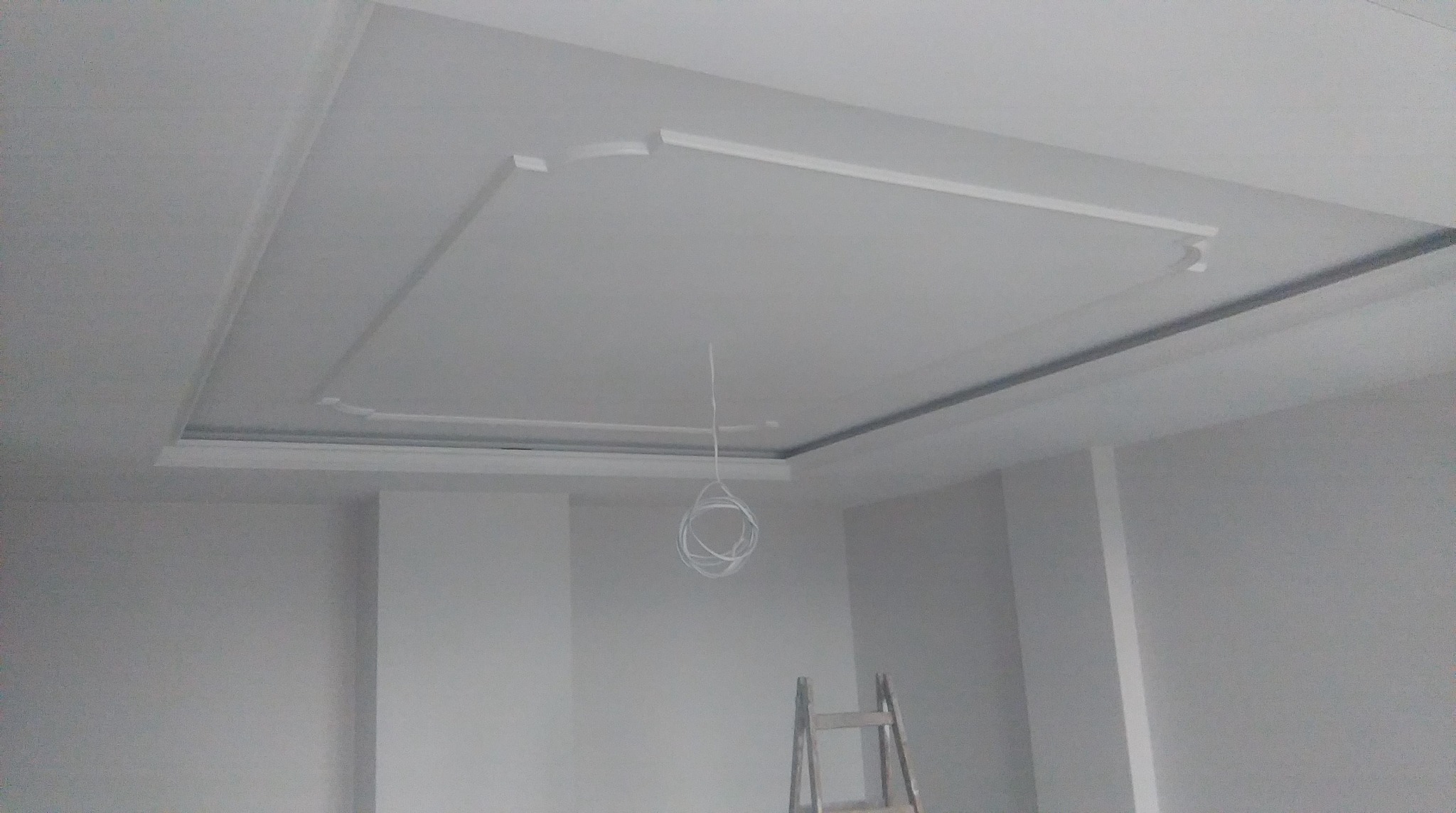Biały sufit z dekoracyjnym, prostokątnym obramowaniem i wystającymi elementami, widoczne kable zwisające z sufitu i drabina w rogu pokoju podczas remontu.