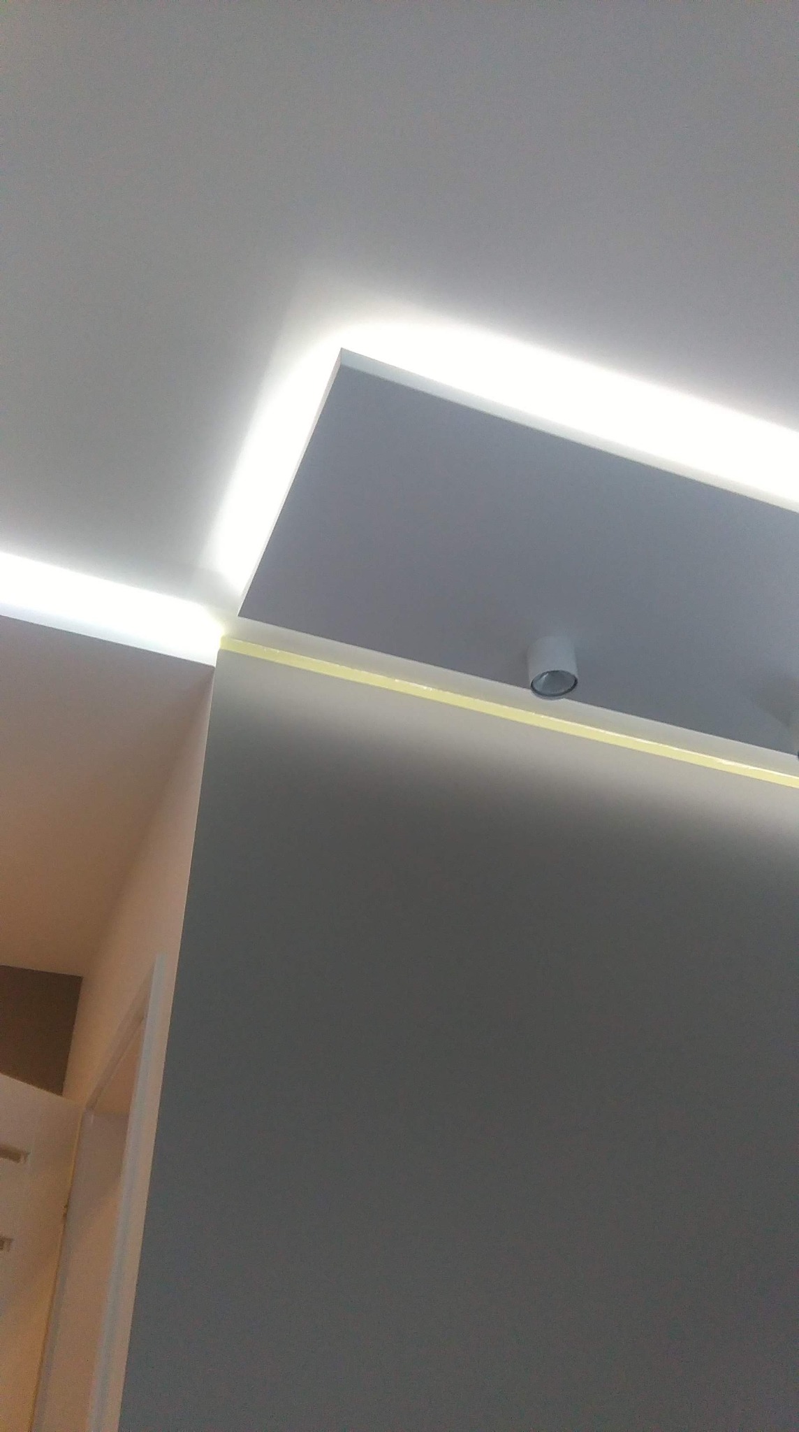 Nowoczesne oświetlenie sufitowe LED w narożniku pokoju, z widoczną taśmą malarską zabezpieczającą ścianę podczas prac wykończeniowych i pojedynczym punktowym reflektorem.