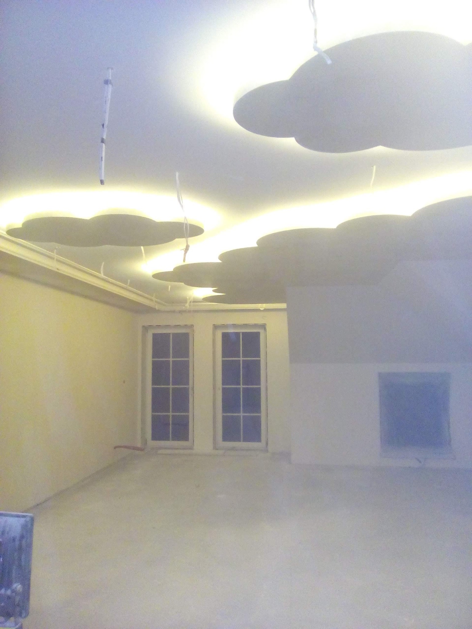 Pokój w trakcie wykończenia z dekoracyjnym oświetleniem sufitowym w kształcie chmur, widoczne okna z białymi ramami i surowa podłoga, ściany w jasnych odcieniach beżu i szarości.