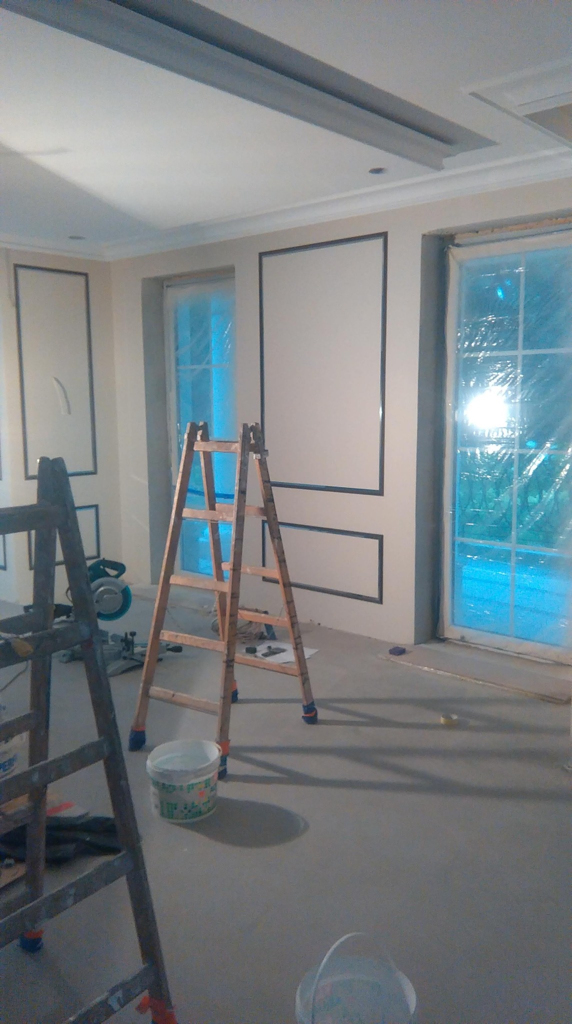 Wnętrze w trakcie prac wykończeniowych: ściany z dekoracyjnymi ramami, okna zabezpieczone folią, drabiny i narzędzia malarskie widoczne na podłodze.