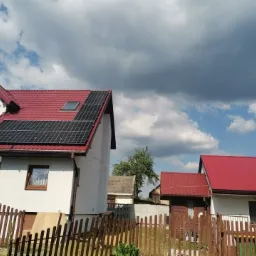 Instalacja fotowoltaiczna na dachu domu z czerwoną dachówką, widok zza drewnianego płotu w słoneczny dzień z chmurami.