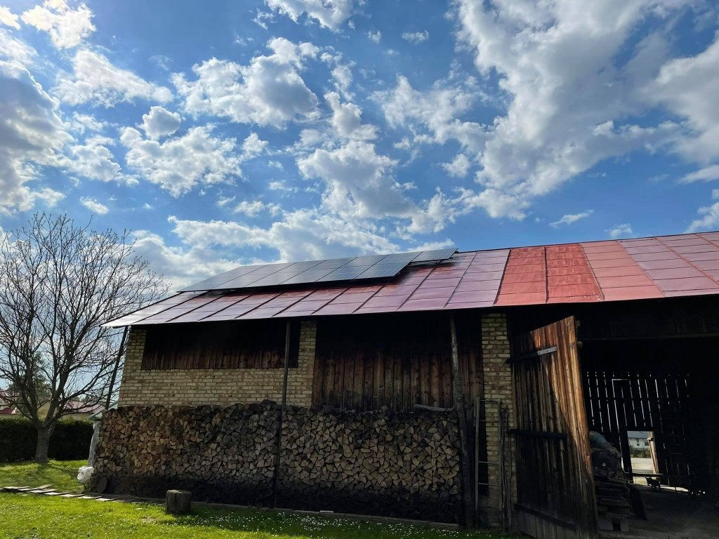 Panele słoneczne zamontowane na dachu stodoły z czerwoną dachówką, składowane drewno na opał przy ścianie, błękitne niebo z obłokami w tle.