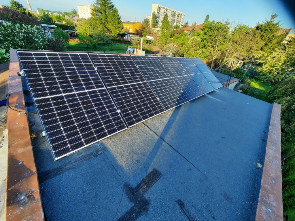 Panele słoneczne zamontowane na płaskim dachu budynku mieszkalnego, widoczne drzewa i budynki w tle.