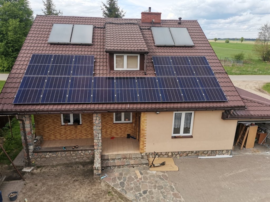 Dom jednorodzinny z brązowym dachem krytym dachówką i panelami fotowoltaicznymi oraz kolektorami słonecznymi na dachu, widok z góry.