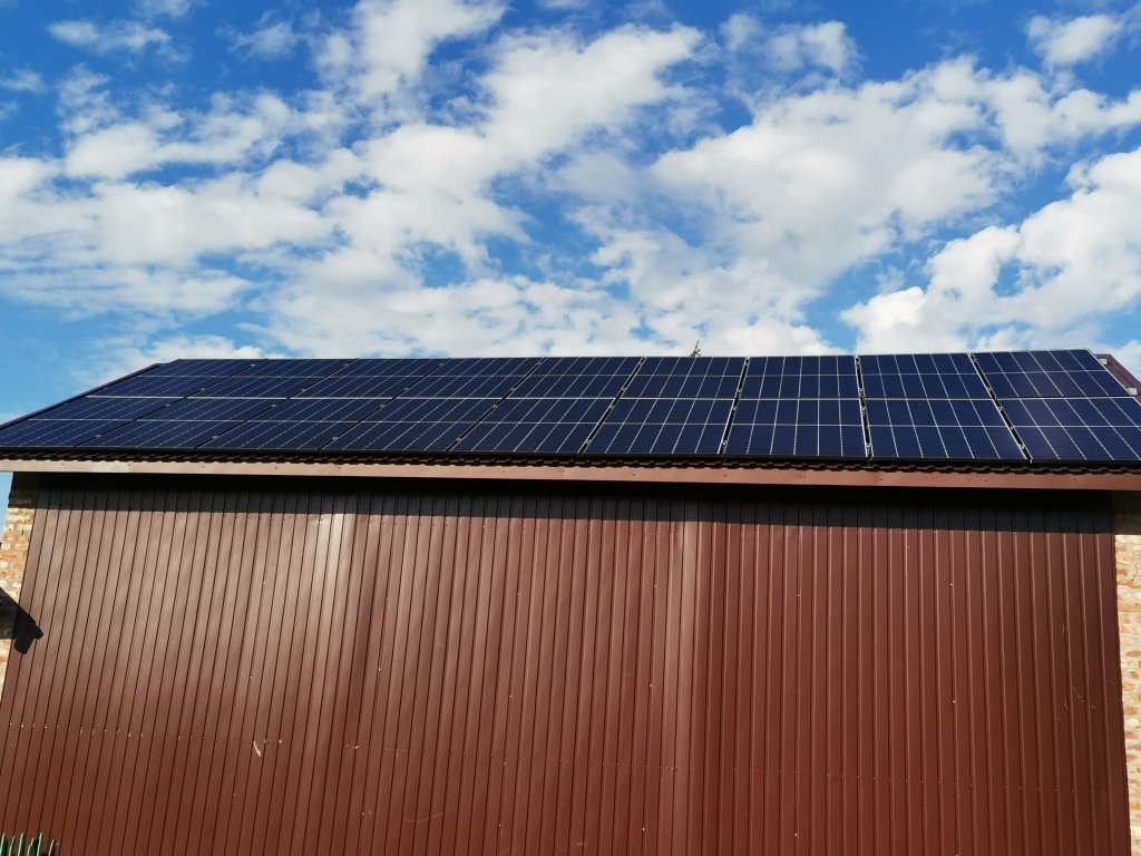 Panele słoneczne na dachu brązowego garażu blaszaka, na tle błękitnego nieba z białymi chmurami.