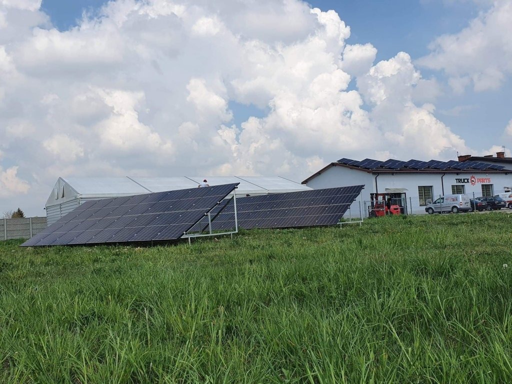 Panele słoneczne na trawiastym terenie przed budynkiem z panelami na dachu, w tle biały namiot i błękitne niebo z chmurami.