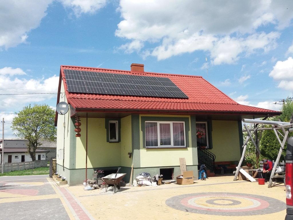 Dom jednorodzinny z panelami słonecznymi na czerwonym dachu, widok z przodu w słoneczny dzień, widoczny wózek budowlany i osoba siedząca na huśtawce.