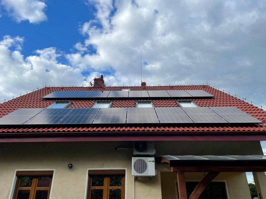 Panele słoneczne na czerwonym dachu domu jednorodzinnego z oknami dachowymi, klimatyzator na ścianie budynku, błękitne niebo z chmurami w tle.