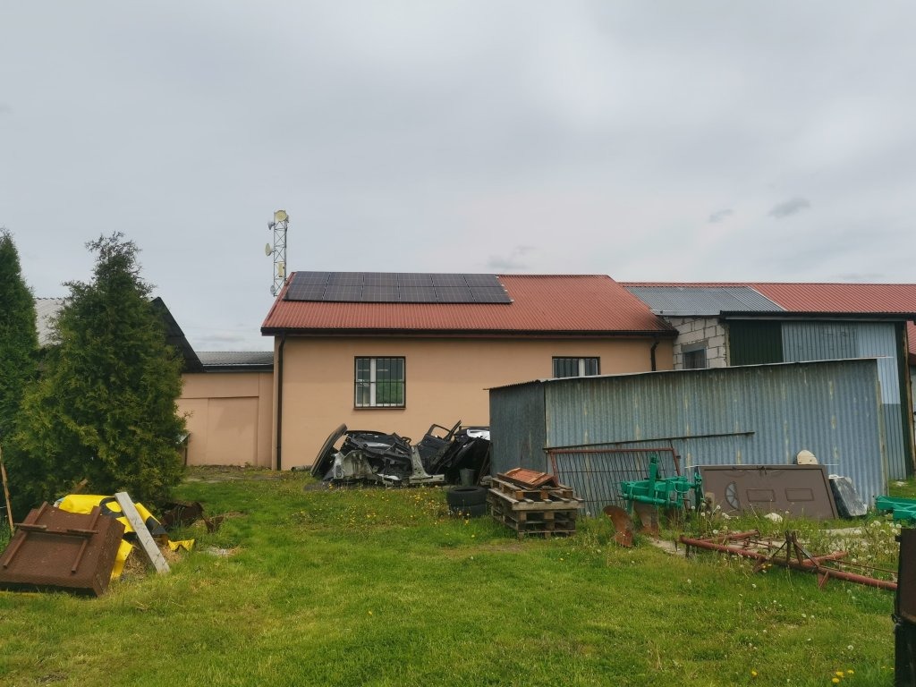 Instalacja paneli słonecznych na dachu budynku gospodarczego z czerwonej dachówki, widok z zewnątrz na tle pochmurnego nieba, z elementami złomu i sprzętu rolniczego na pierwszym planie.