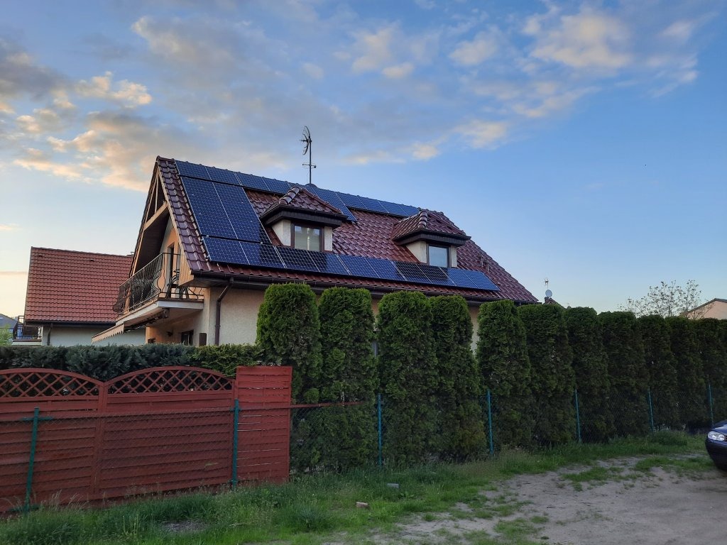 Dom jednorodzinny z panelami fotowoltaicznymi na dachu, widok z poziomu ulicy, ogrodzenie z brązowych desek i wysokie tuje wzdłuż posesji.