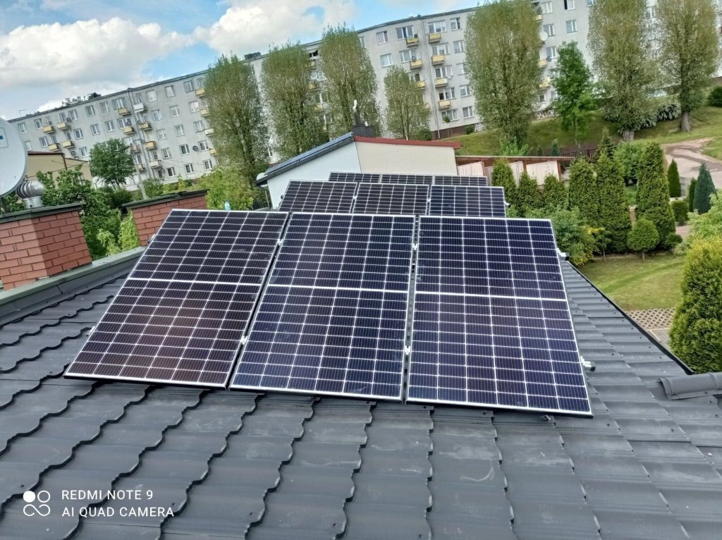 Panele słoneczne zamontowane na ciemnoszarym dachu z blachodachówki modułowej, w tle budynek mieszkalny i drzewa.