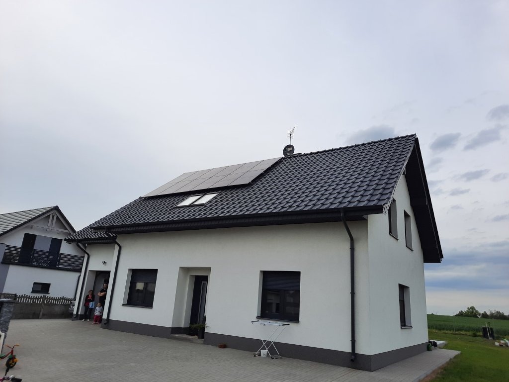 Biały dom jednorodzinny z czarnym dachem i panelami słonecznymi, widok z zewnątrz, na podjeździe stoi rodzina.