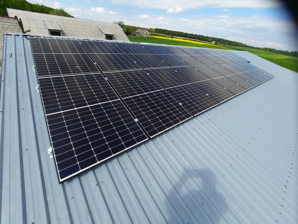 Panele słoneczne zamontowane na szarym, blaszanym dachu budynku gospodarczego z widokiem na pola i błękitne niebo.