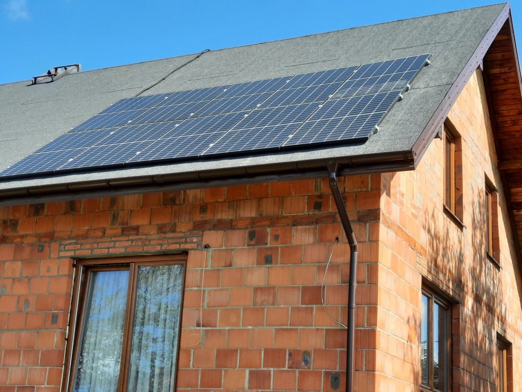 Panele słoneczne zamontowane na dachu domu z czerwonej cegły, widoczne okna i rynna spustowa, błękitne niebo w tle.