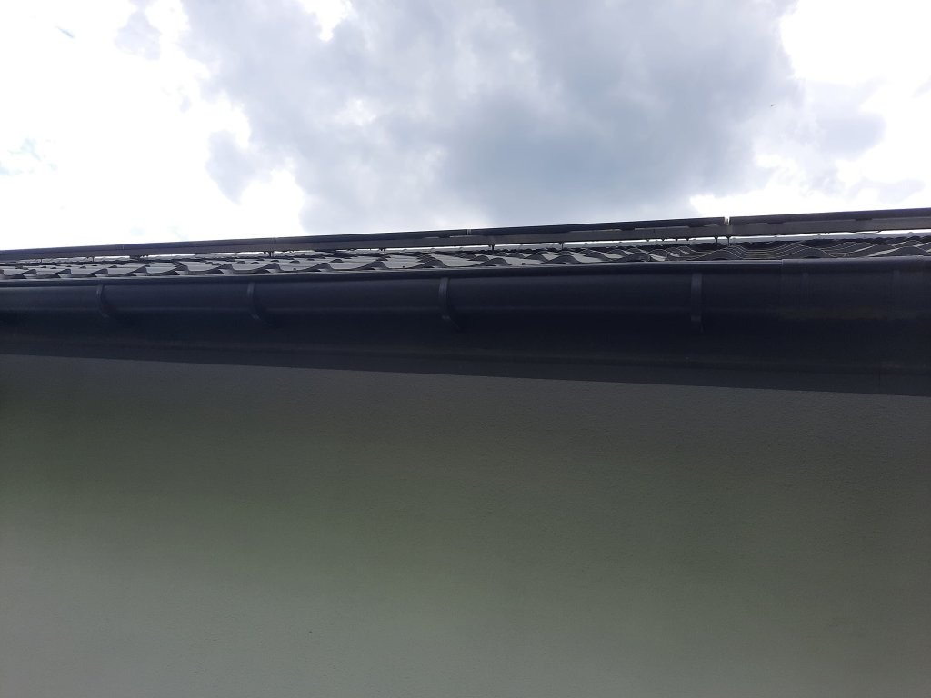 Fragment dachu z czarną dachówką i systemem rynnowym na tle pochmurnego nieba.