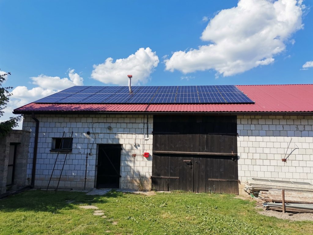 Instalacja paneli słonecznych na dachu budynku gospodarczego z białej cegły, częściowo pokrytego czerwoną blachą, widok w słoneczny dzień.