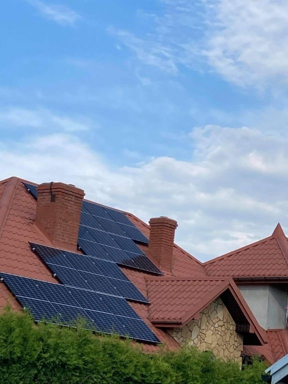 Panele słoneczne na dachu domu z czerwoną dachówką i ceglanym kominem, widok z dołu na tle błękitnego nieba z chmurami.