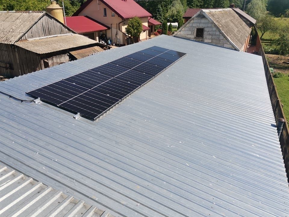 Panel słoneczny zamontowany na blaszanym dachu budynku gospodarczego z widocznymi budynkami mieszkalnymi w tle.
