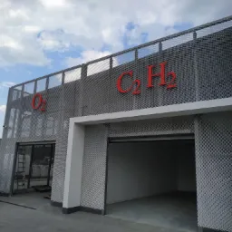 Nowoczesny budynek przemysłowy z metalową elewacją i czerwonymi oznaczeniami chemicznymi C2H2 i O2, widoczny biały samochód dostawczy i sprzęt budowlany w tle.