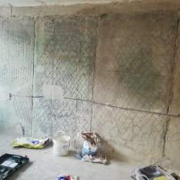 Mad-Wyk - Przygotowana do położenia płytek ściana z siatką nacięć, widoczne worki z zaprawą, wiadro z klejem i poziomica.