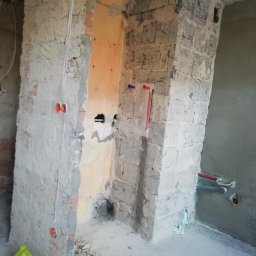 Mad-Wyk - Wnętrze w trakcie prac przygotowawczych do remontu łazienki; widoczne fragmenty cegieł, instalacja elektryczna, rury wodociągowe oraz resztki gruzu na podłodze, żółty kanister z płynem w rogu.