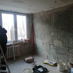 Mad-Wyk - Wnętrze w trakcie remontu: ściany przygotowane pod tynk z siatką, okno z grzejnikiem, drabina malarska, wiadro z resztkami farby i narzędzia na podłodze.