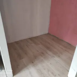 Wnętrze pomieszczenia w trakcie prac wykończeniowych, widoczna podłoga z paneli imitujących drewno, ściany częściowo pokryte gładzią, jedna ściana w kolorze różowym, w rogu przy futrynie drzwiowej...