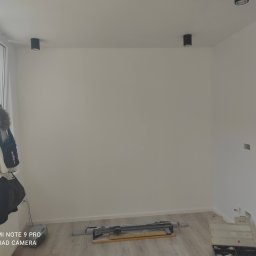 Mad-Wyk - Pomalowane na biało pomieszczenie z dwoma czarnymi lampami sufitowymi, narzędziem do cięcia płytek na podłodze i kurtką wiszącą na grzejniku przy oknie. Świeżo położone panele podłogowe.