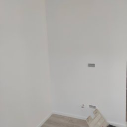 Mad-Wyk - Świeżo pomalowane na biało ściany w narożniku pomieszczenia, z widocznymi puszkami instalacyjnymi, listwami przypodłogowymi i rozłożonymi narzędziami w kuwecie na podłodze imitującej drewno.