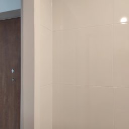 Mad-Wyk - Fragment wykończonej łazienki z beżowymi, błyszczącymi płytkami ceramicznymi na ścianie, widoczny fragment brązowych drzwi wejściowych z wizjerem i biała ościeżnica.