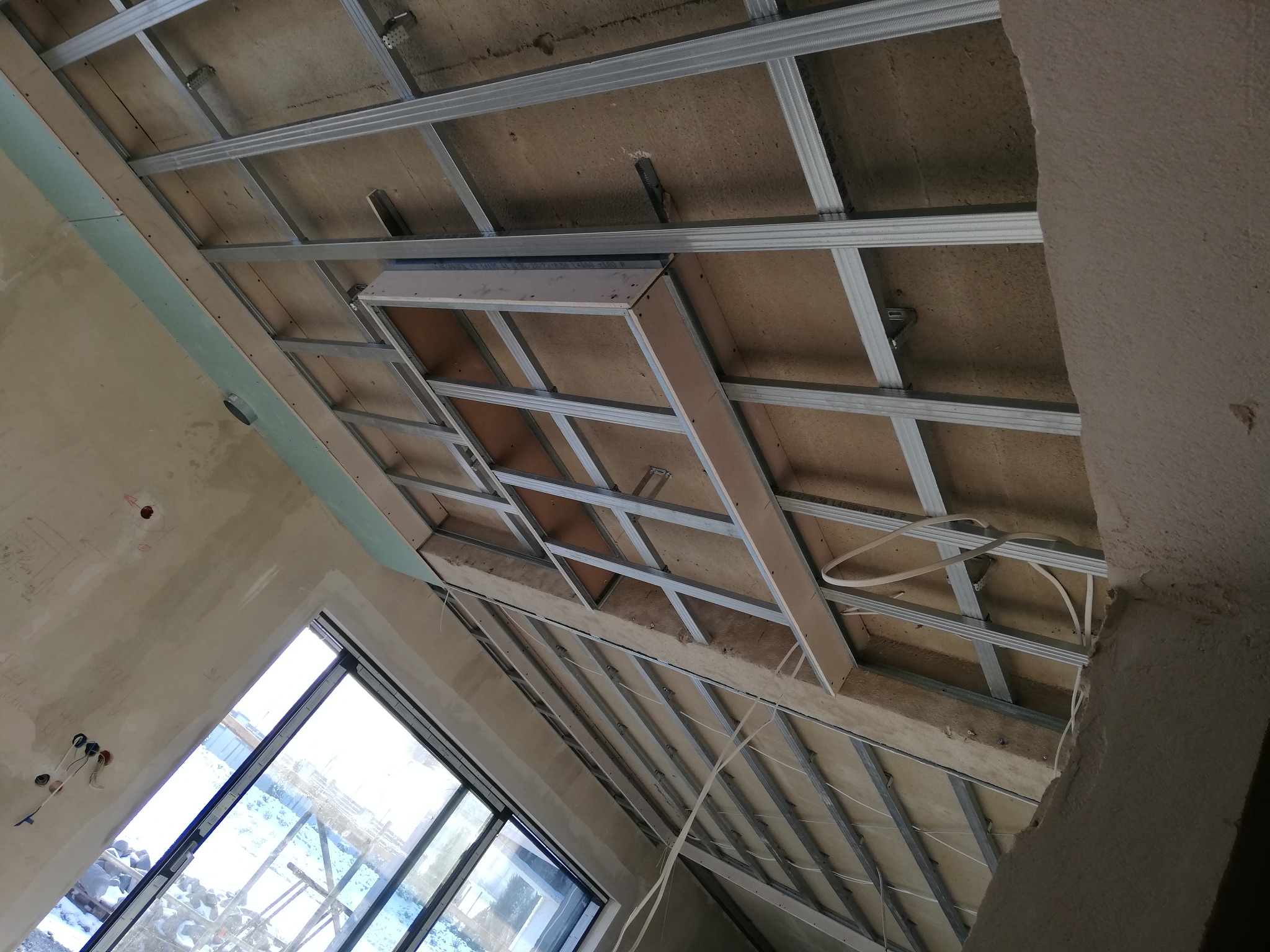 Sufit podwieszany z metalową konstrukcją szkieletową i częściowo zamontowanymi płytami, widok z dołu pod kątem, widoczne przewody elektryczne i okno.