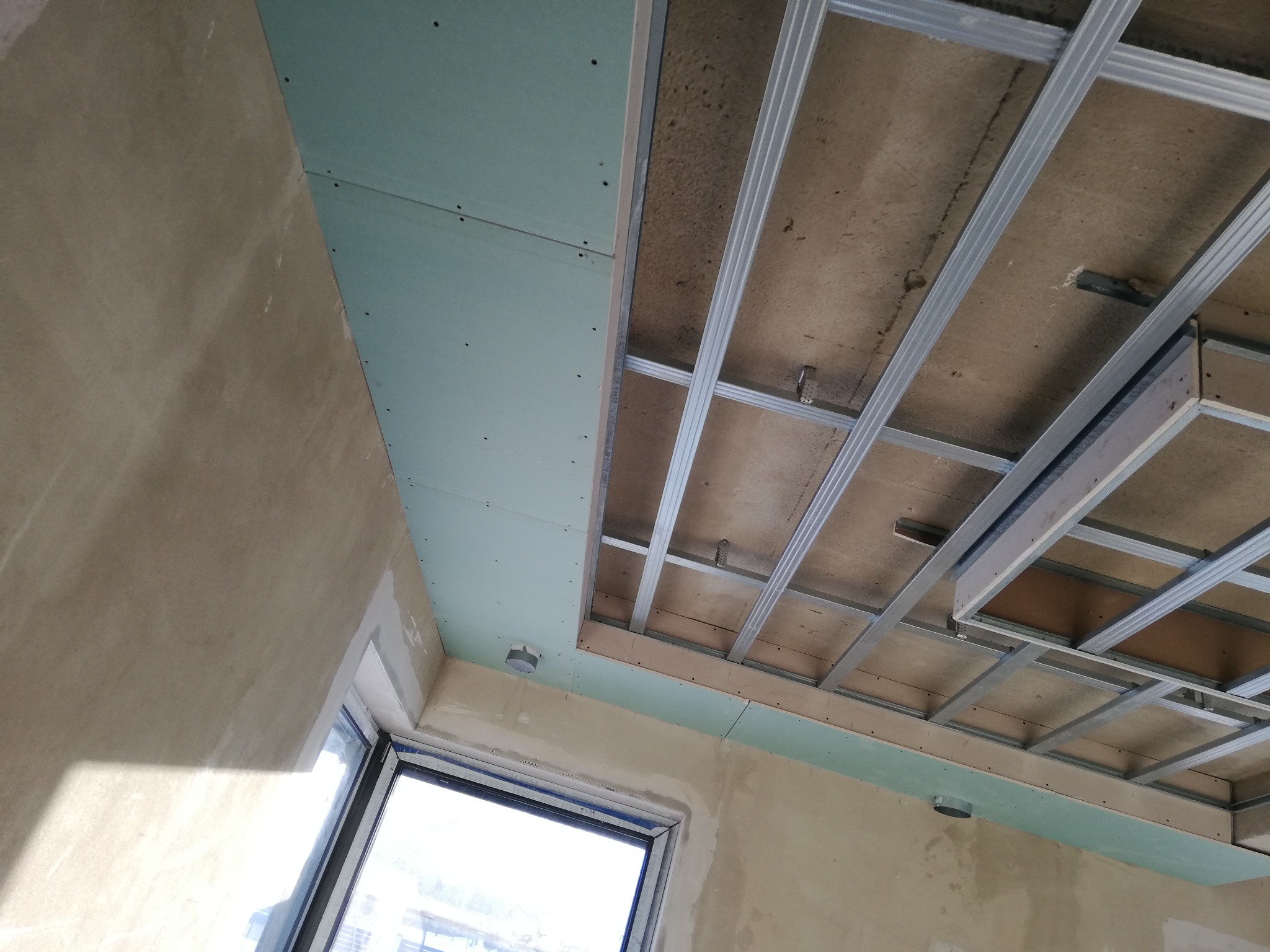 Montaż sufitu podwieszanego z płyt karton-gips: widoczne metalowe profile konstrukcyjne, częściowo zamontowane zielone płyty G-K, surowy betonowy strop powyżej i fragment okna.