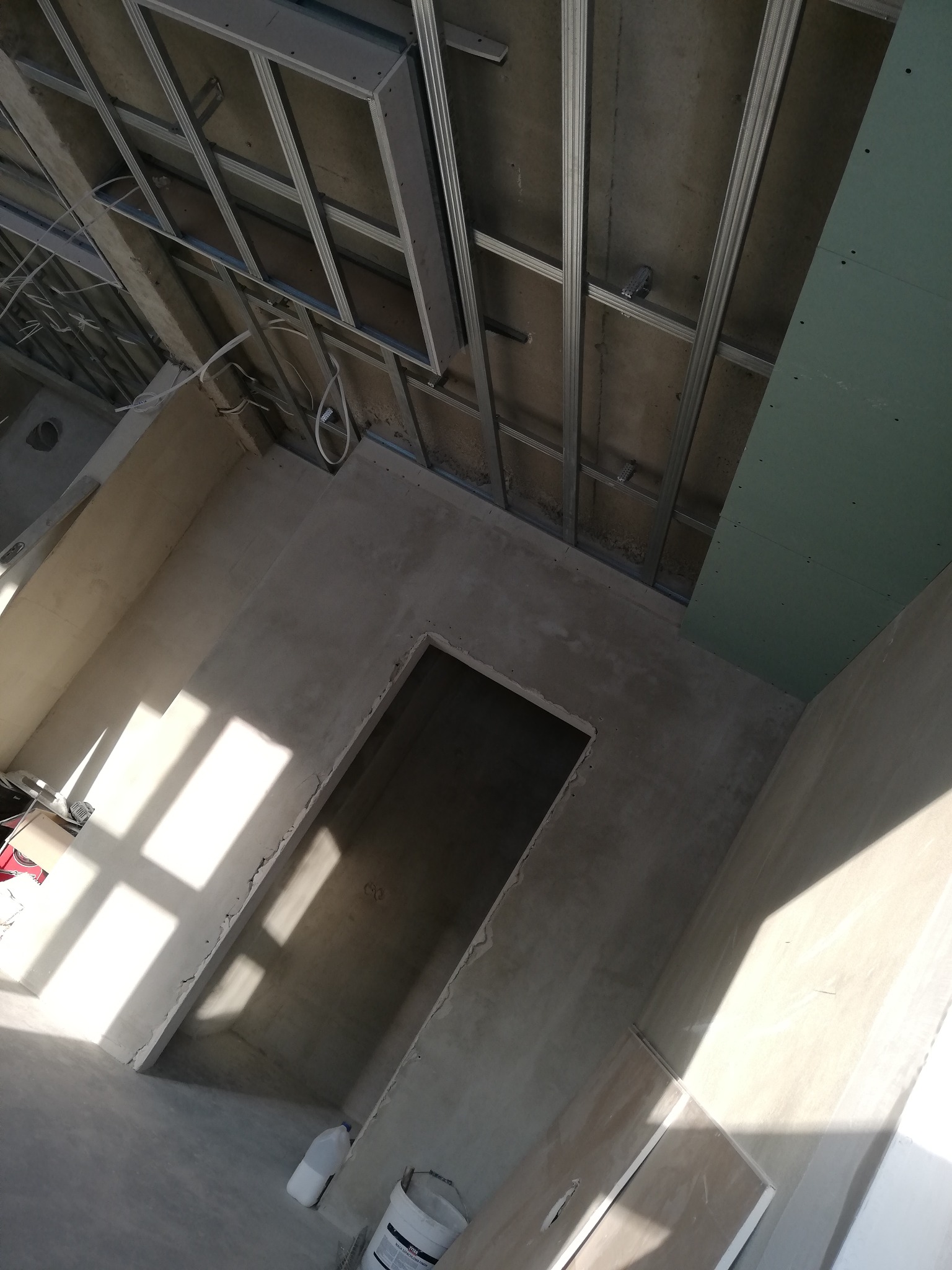 Wnętrze w trakcie prac wykończeniowych: widoczne profile sufitowe, częściowo zamontowana płyta gipsowo-kartonowa, ściany po nałożeniu gładzi, wpadające promienie słoneczne przez otwór okienny.