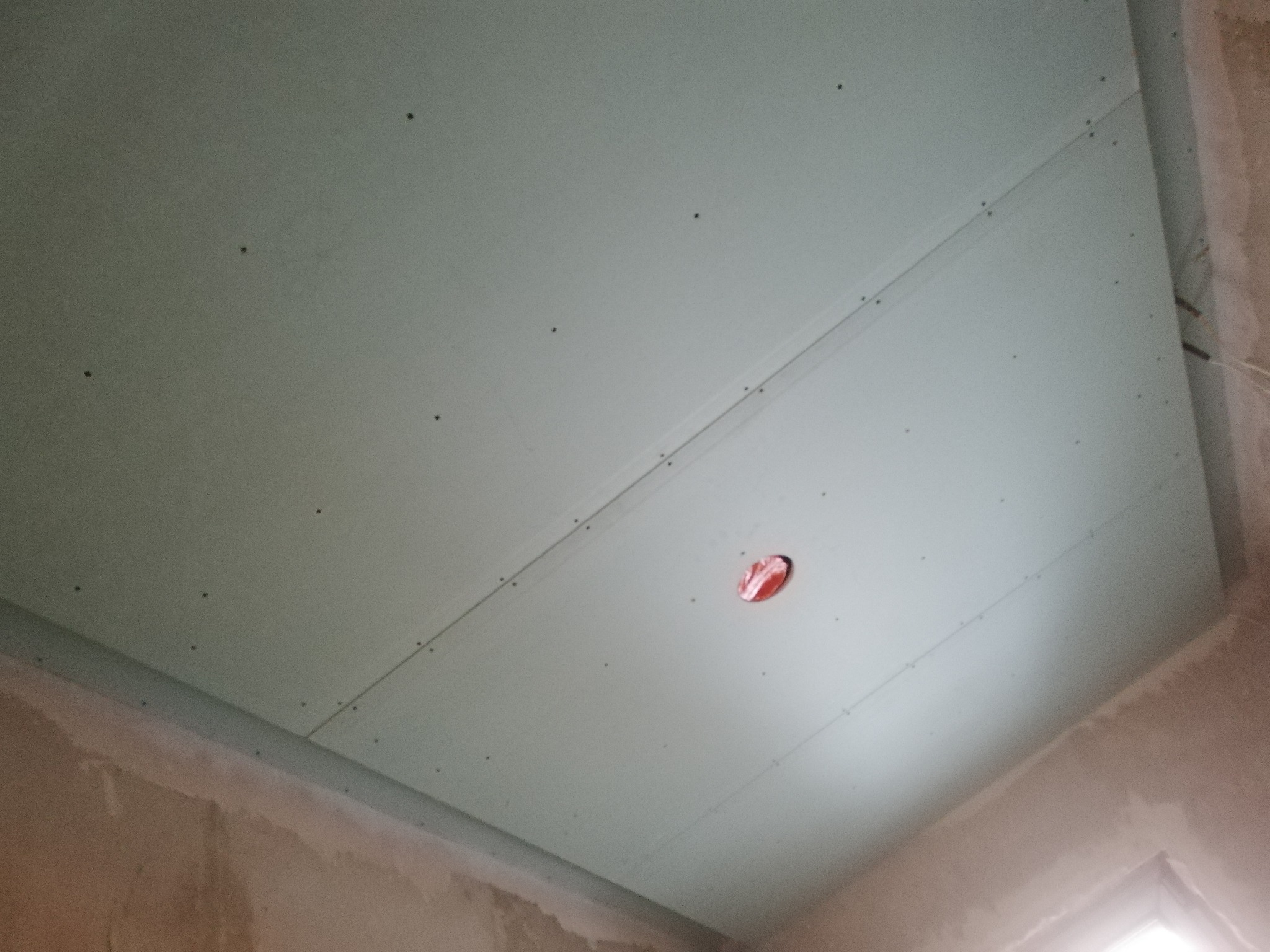 Sufit z zamontowaną płytą gipsowo-kartonową, widoczne ślady po wkrętach i otwór na instalację elektryczną, surowe ściany w tle.