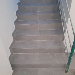 Schody wyłożone szarymi, matowymi płytkami gresowymi, widok z góry, częściowo widoczna metalowa barierka ochronna.