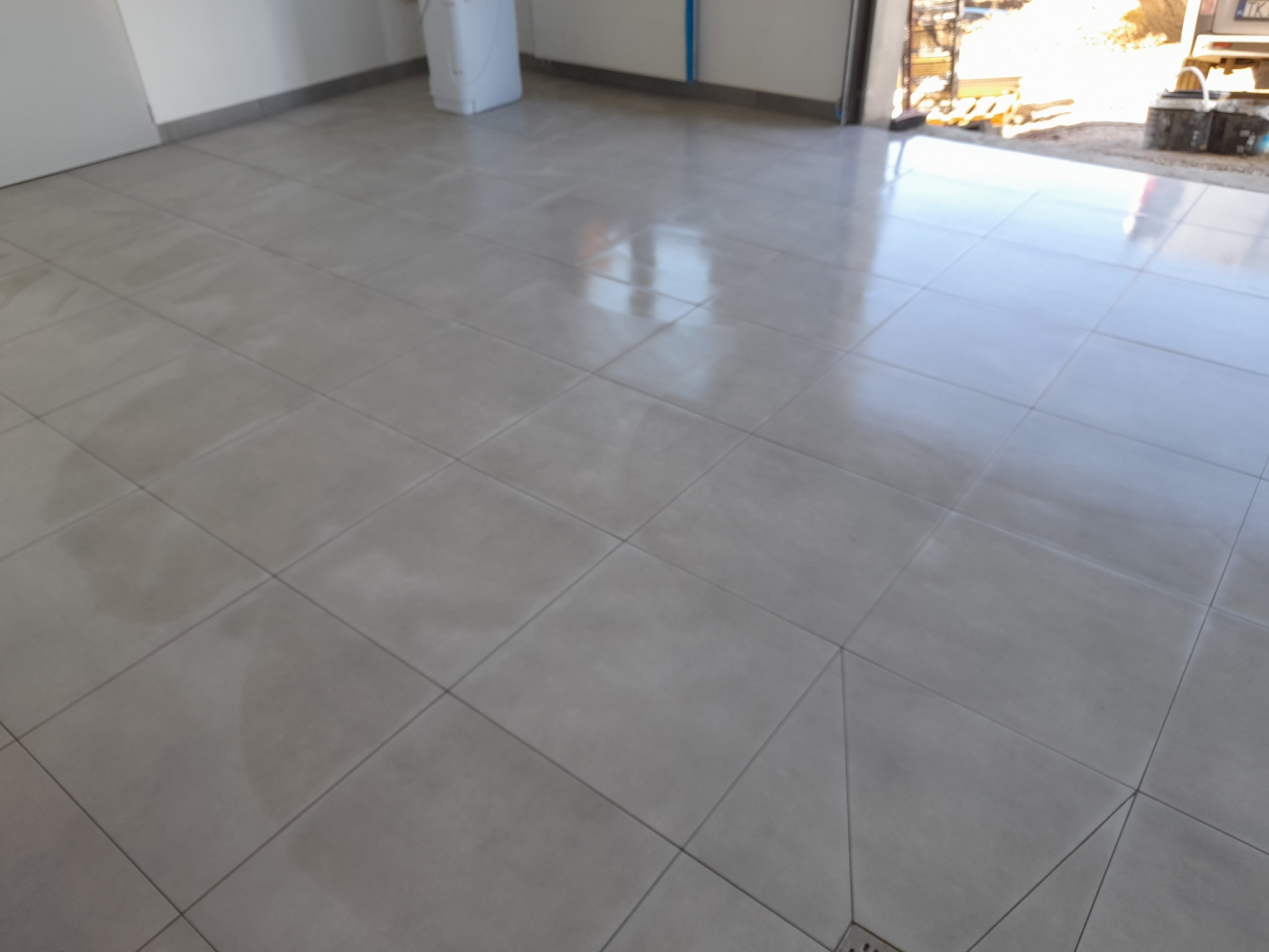 Ułożone szare płytki podłogowe w garażu, widoczny odpływ liniowy, w tle otwarte drzwi garażowe z widocznymi elementami konstrukcyjnymi i podjazdem.