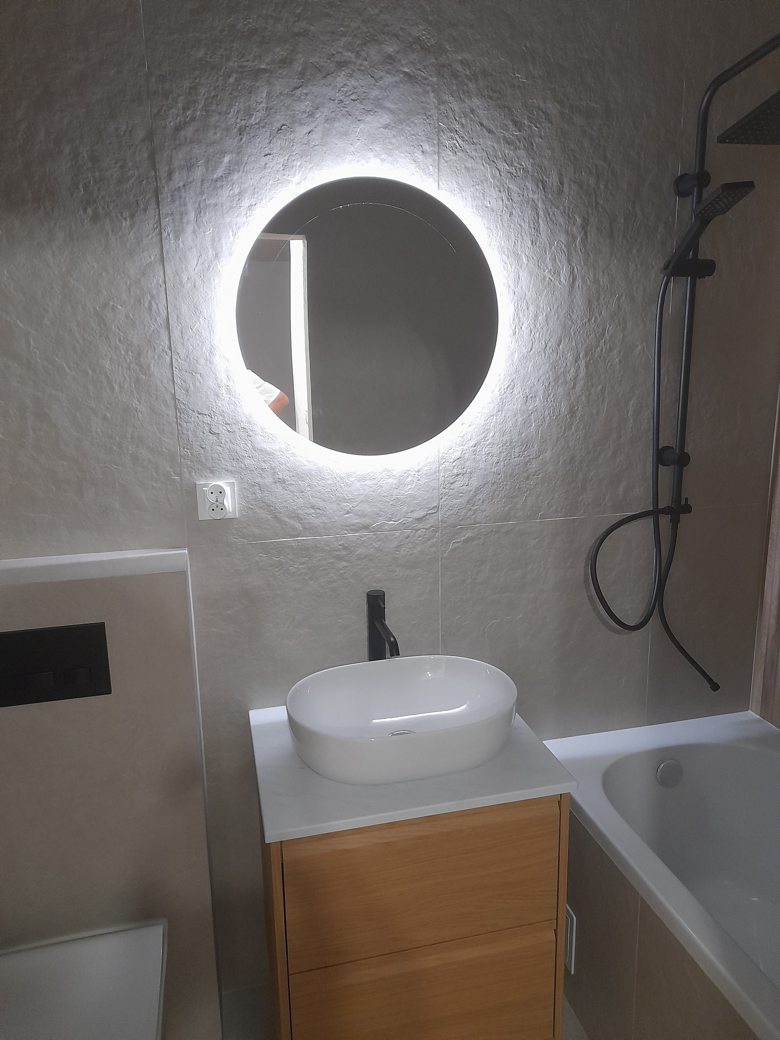 Nowoczesna łazienka z okrągłym lustrem podświetlanym LED, umywalką nablatową na drewnianej szafce, szarą płytką imitującą kamień na ścianie i czarną armaturą.