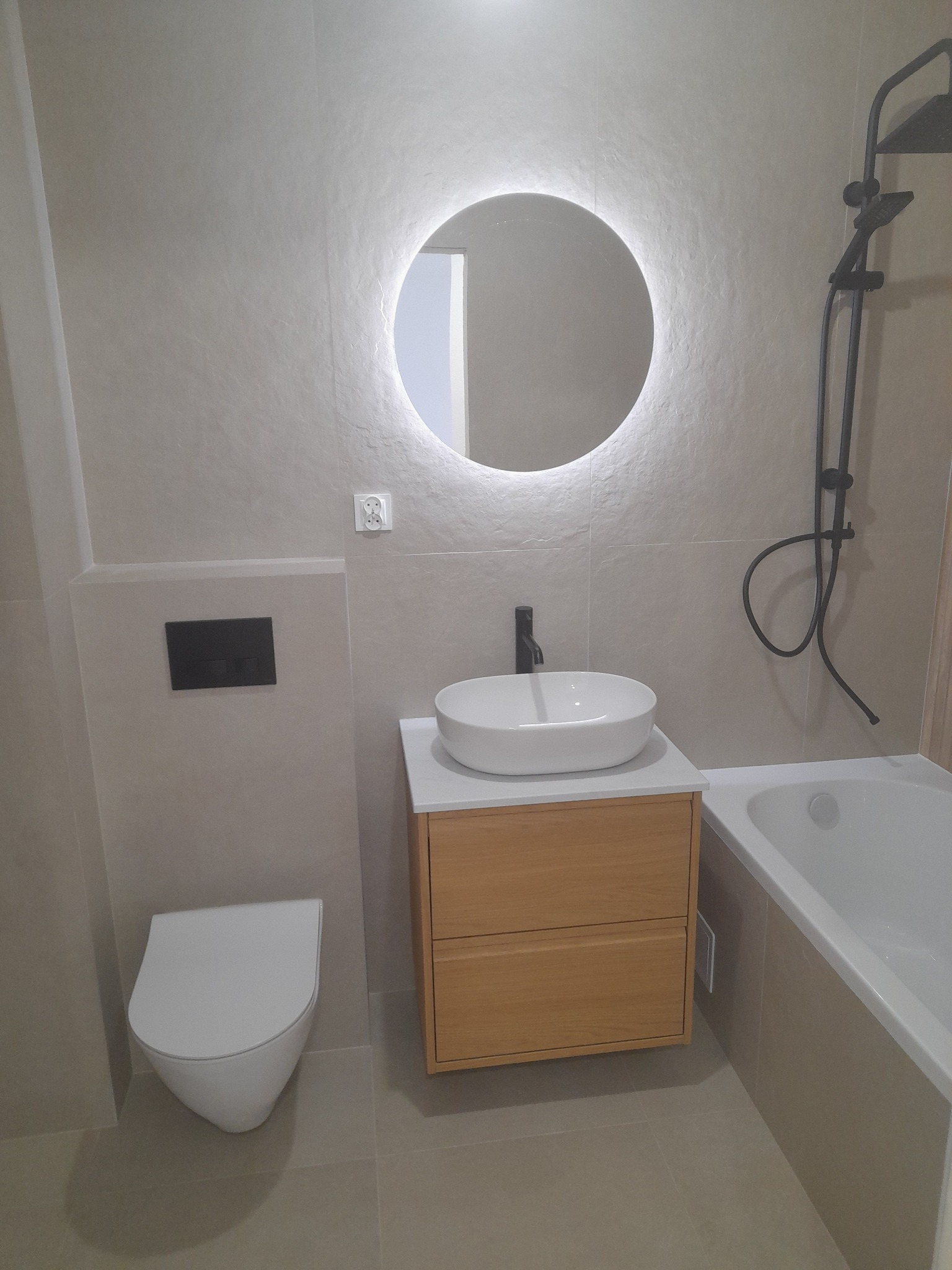 Nowoczesna łazienka po remoncie: beżowe płytki, podwieszana toaleta z czarnym przyciskiem, umywalka nablatowa na drewnianej szafce z okrągłym, podświetlanym lustrem i czarną armaturą oraz czarny...