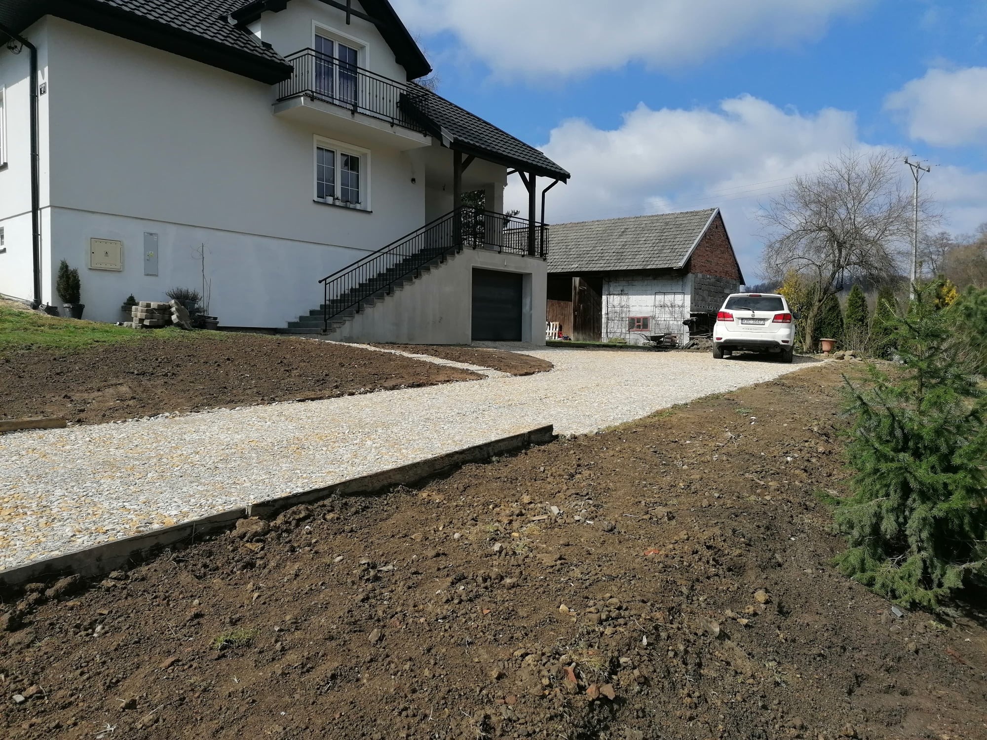 Podjazd do domu z jasnym żwirem, obrzeża betonowe, świeżo przekopana ziemia, biały dom z balkonem i garażem w tle, samochód zaparkowany obok budynku gospodarczego.