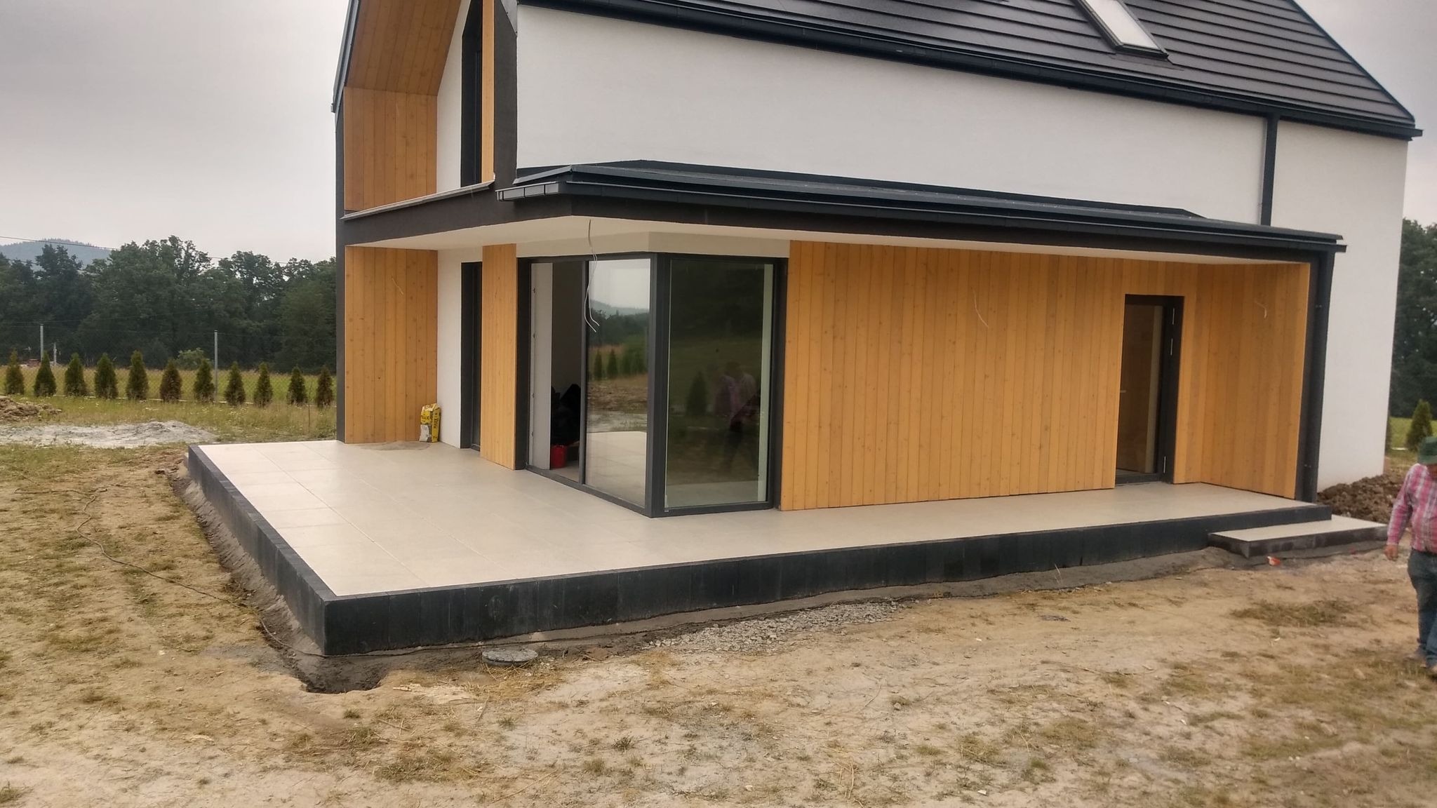 Nowoczesny taras wyłożony jasnymi płytami ceramicznymi, obrzeże z ciemnej kostki, drewniana elewacja domu w tle, widoczne przygotowania terenu.