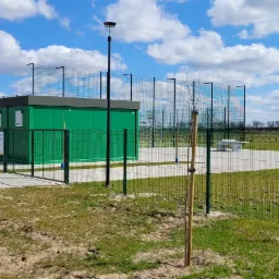 Nowo wybudowany, zielony kontener sanitarny z oświetleniem zewnętrznym i ogrodzeniem z siatki na terenie rekreacyjnym z boiskami sportowymi w tle, słoneczny dzień.