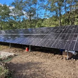 Panel słoneczny zamontowany na metalowej konstrukcji w terenie z drzewami w tle, widoczne pudełka i zwój kabla.