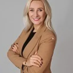 Portret uśmiechniętej blondynki w brązowym żakiecie, z założonymi rękami, na jasnoszarym tle. Kadr od pasa w górę.