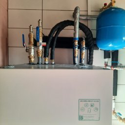 NIKIEL INSTALACJE SANITARNE, C.O., GAZ - Instalacja hydrauliczna z niebieskim zbiornikiem wyrównawczym i licznymi zaworami oraz rurami w izolacji, przyłącza do urządzenia Aquarea Smart Cloud firmy Panasonic na tle białych płytek.