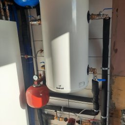 NIKIEL INSTALACJE SANITARNE, C.O., GAZ - Biały zasobnik ciepłej wody Galmet z podłączonymi instalacjami hydraulicznymi, w tym czerwony zbiornik przeponowy i rury w izolacji, zamontowany na ścianie wyłożonej białymi płytkami.