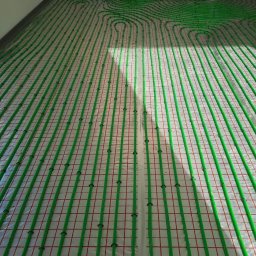 NIKIEL INSTALACJE SANITARNE, C.O., GAZ - Zielone rury ogrzewania podłogowego ułożone na folii z czerwoną siatką, widok z góry, w tle biała ściana i schody.