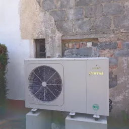 Zewnętrzna jednostka pompy ciepła marki Galmet Prima, zamontowana na betonowych podstawach przy nier&oacute;wnej, ceglano-tynkowanej ścianie budynku z zakratowanym okienkiem.