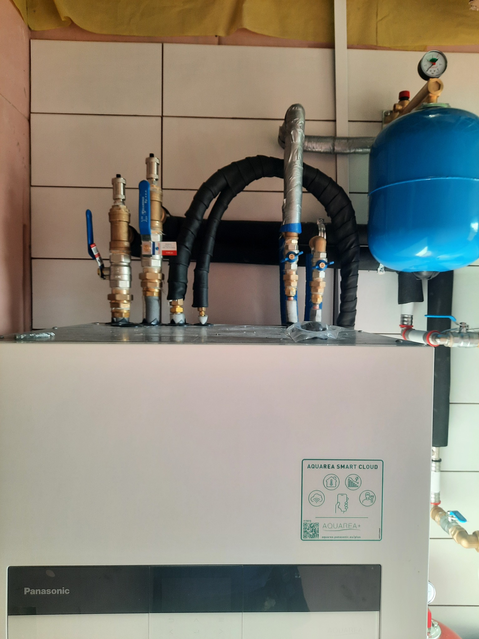 Instalacja hydrauliczna z niebieskim zbiornikiem wyrównawczym i licznymi zaworami oraz rurami w izolacji, przyłącza do urządzenia Aquarea Smart Cloud firmy Panasonic na tle białych płytek.