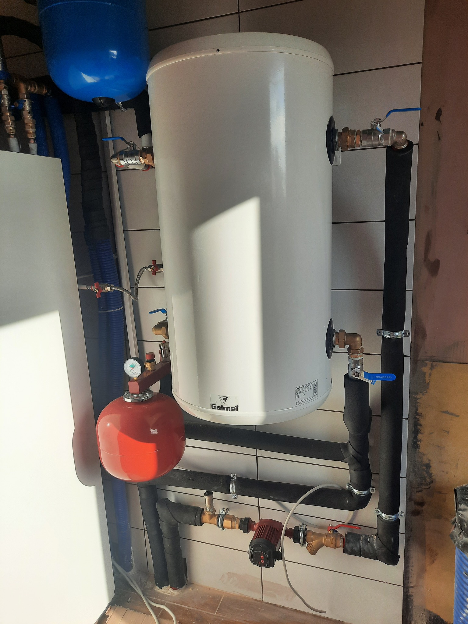 Biały zasobnik ciepłej wody Galmet z podłączonymi instalacjami hydraulicznymi, w tym czerwony zbiornik przeponowy i rury w izolacji, zamontowany na ścianie wyłożonej białymi płytkami.